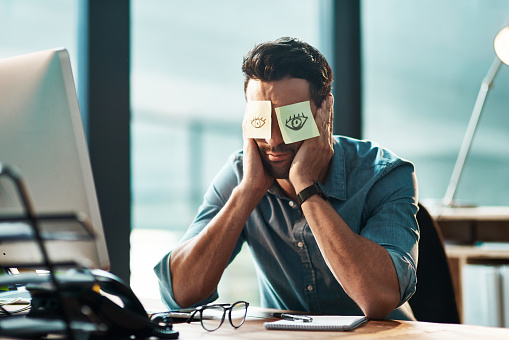 Shot of a tired young businessman working at his desk with adhesive notes covering his eyes