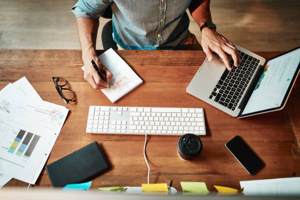 Hands that were made to work hard Cropped high angle shot of a businessman using a laptop and writing notes at his desk in a modern office multitasking stock pictures, royalty-free photos & images