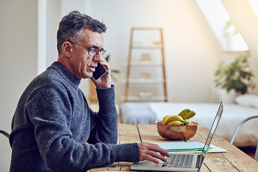 Shot of a mature man on a call while using a laptop at home