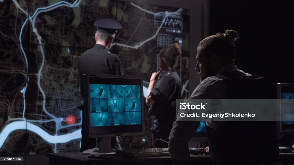 Policemen chasing gang from surveillance office Professional policemen tracing group of bandits in room of video monitoring watching movements on city map while other colleagues working Military Stock Photo