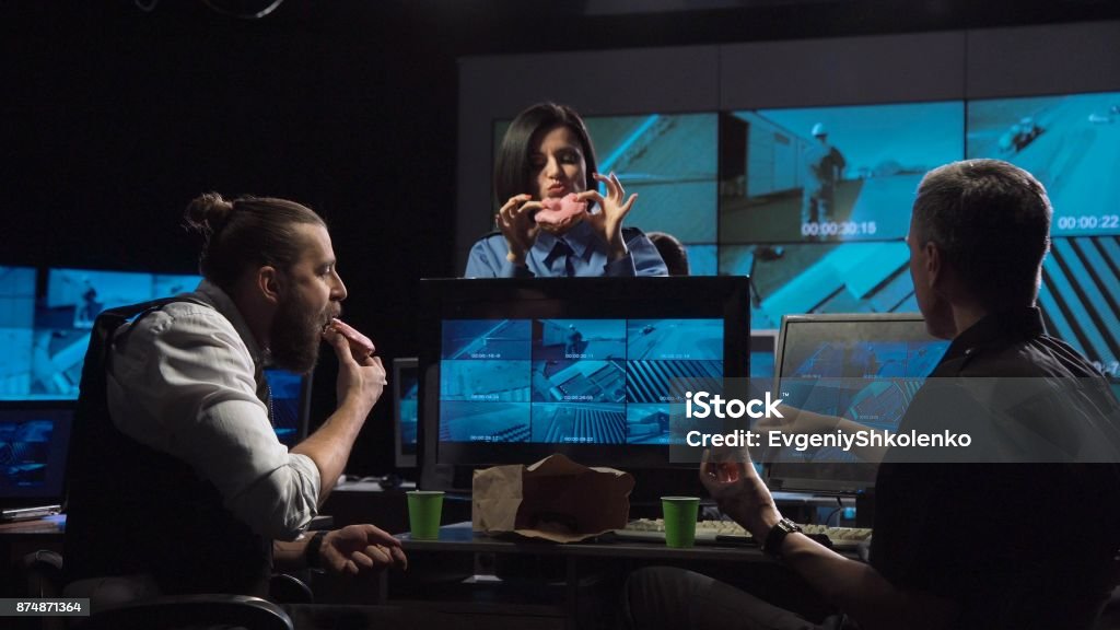 Security guards watching the surveillance cameras Security guards eating donuts while watching the surveillance cameras on screen while working late at night in an office with modern equipment Eating Stock Photo