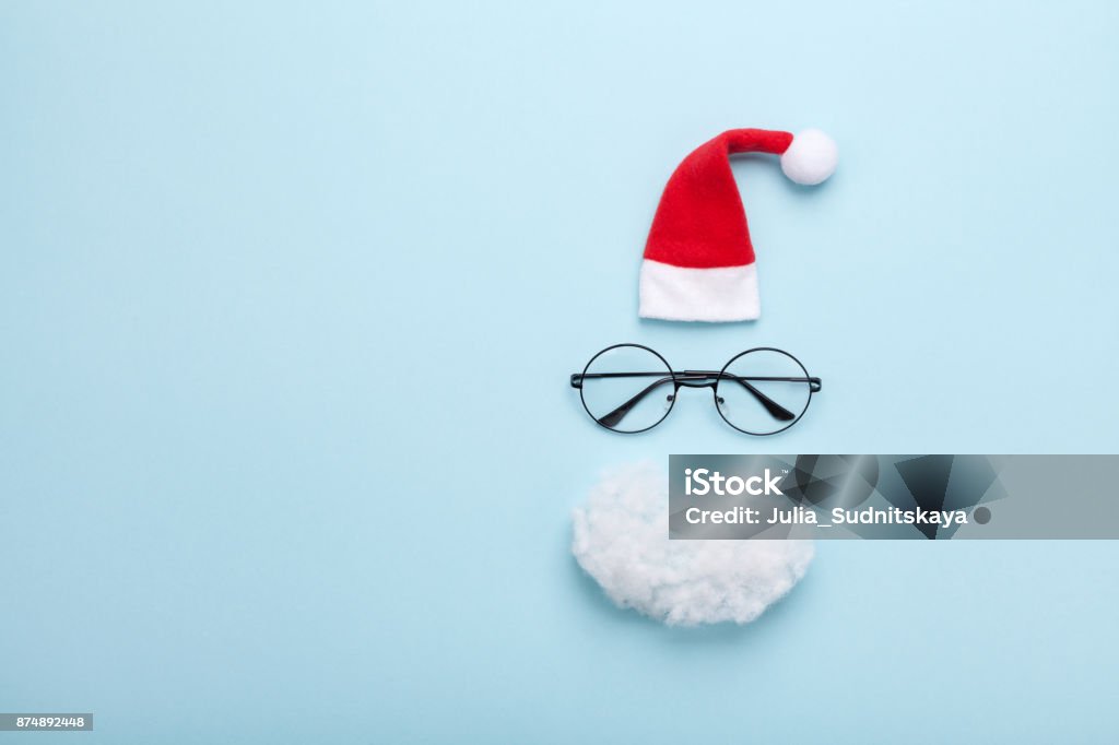 Creative christmas composition. Greeting card, invitation or flyer. Santa hat, beard and glasses on blue background top view. Creative christmas composition. Greeting card, invitation or flyer. Santa hat, beard and glasses on blue background top view. Flat lay. Copy space for text and design. Eyeglasses Stock Photo