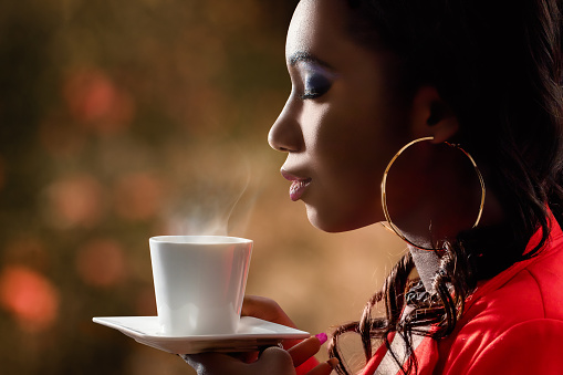 Close up side view of attractive african woman smelling scent of hot beverage.Low key portrait of woman with directional backlit ambient light holding cup with coffee.