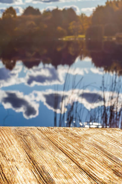 istock Empty rustic wood table top on  blurred colorful autumn landscape background with fall forest and blue sky with cloudscape reflection on the lake. Can montage or display your products 874962792