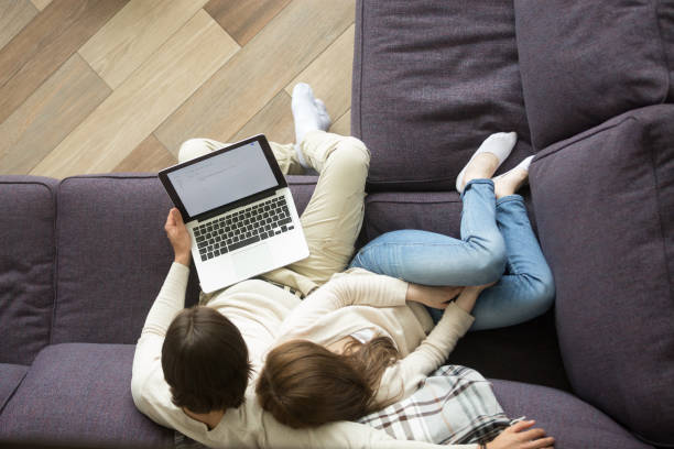 istock Young couple sitting on couch embracing holding laptop, top view 874986374
