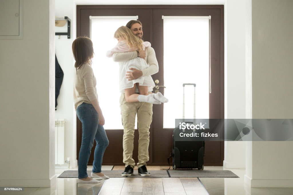 Father arrived came home returning after trip holding hugging daughter Happy father arrived home returning after business trip with baggage, daddy missed little daughter holding in arms hugging girl while wife standing in hall, family reunion, welcome back dad concept Arrival Stock Photo