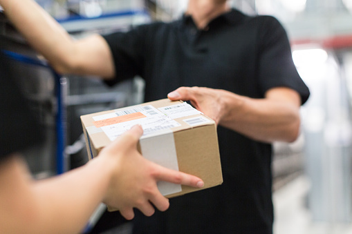 Male warehouse worker giving a box to female coworker. Focus on cardboard box and hands.