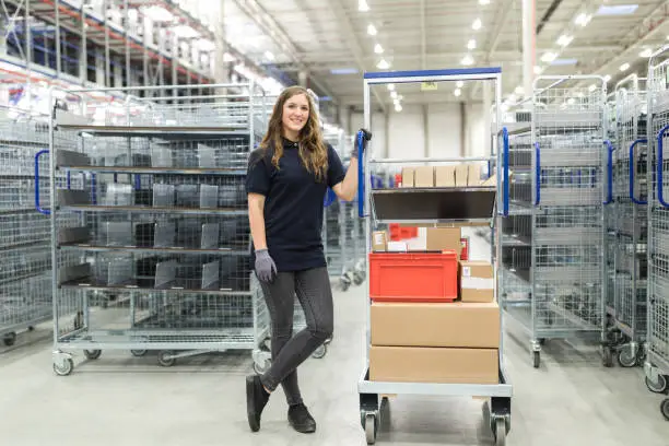 Happy female distribution warehouse worker standing by trolley in plant. Woman in uniform moving a rack with boxes in warehouse.