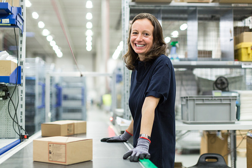 Portrait of smiling young woman working in large distribution warehouse standing by conveyor belt.