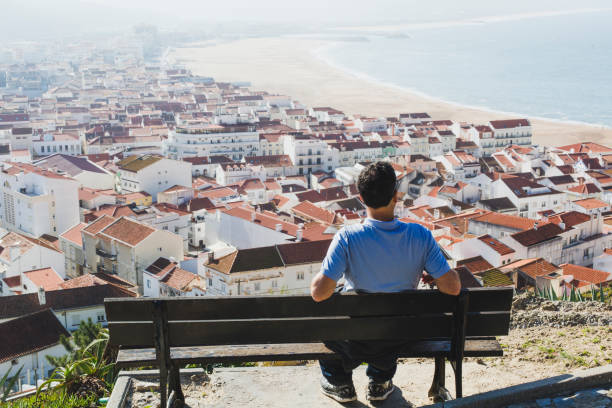 mature man sitting on a bench and looking at nazare town and beach, portugal - sky sea town looking at view imagens e fotografias de stock