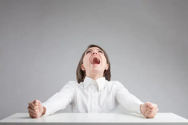 high pressure with anger and stress concept, in a portrait of a woman on a gray background that's shouting to the sky with her punches clenched