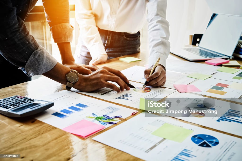 Young businessmen and partnership holding a pen pointing the graph to analyze the marketing plan with calculater and laptop computer on wood desk in office. Improvement Stock Photo