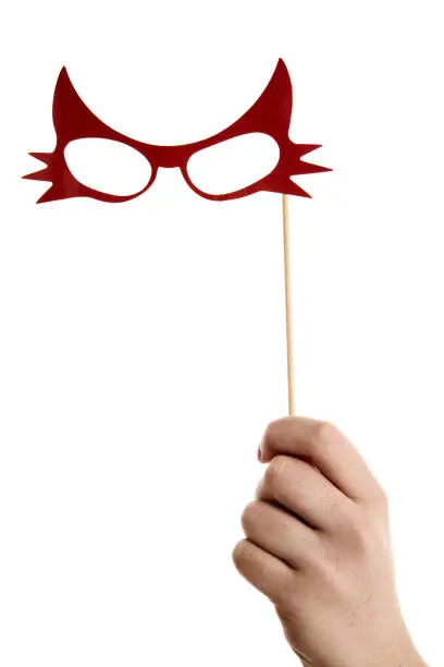A woman's hand holds up a photo booth prop in the shape of catwoman spectacles against awhite background.