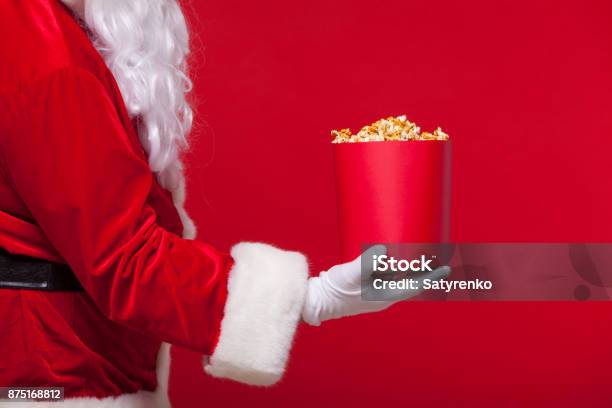 Christmas Photo Of Santa Claus Gloved Hand With A Red Bucket With Popcorn On A Red Background Stock Photo - Download Image Now