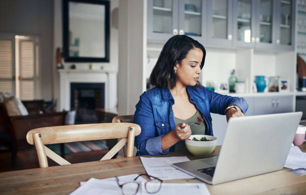 Managing her time like a pro Shot of a young woman checking her watch and having a salad while working from home checking the time stock pictures, royalty-free photos & images