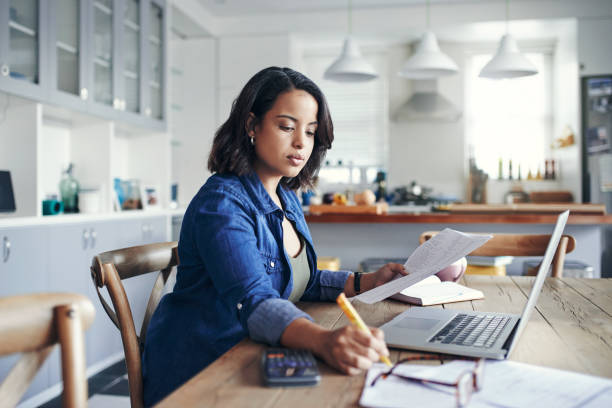 Getting her home business up and running Shot of a young woman using a laptop and  going through paperwork while working from home budget stock pictures, royalty-free photos & images