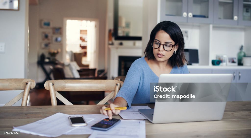 What's the budget looking like this month? Shot of a young woman using a laptop and calculator while working from home Calculator Stock Photo