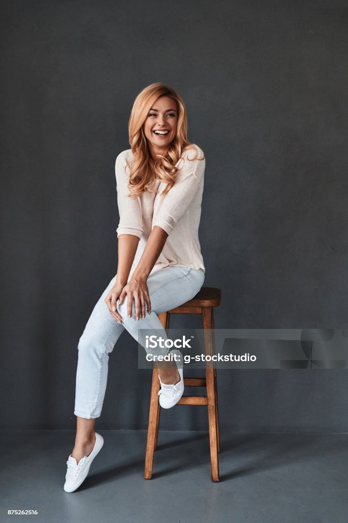 Proud to be beautiful. Full length studio shot of attractive young woman looking at camera and smiling while sitting on stool against grey background Women Stock Photo