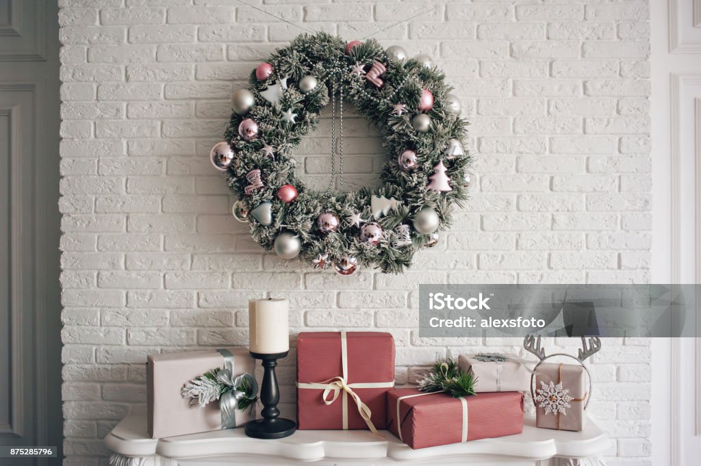 A traditional bright Christmas wreath hanging over the fireplace, on a white brick wall, and packaged gifts are stacked on a fireplace with candles. A traditional bright Christmas wreath hanging over the fireplace, on a white brick wall, and packaged gifts are stacked on a fireplace with candles. Christmas concept, new year. Christmas Stock Photo