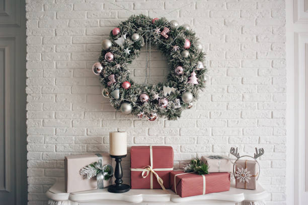 A traditional bright Christmas wreath hanging over the fireplace, on a white brick wall, and packaged gifts are stacked on a fireplace with candles. A traditional bright Christmas wreath hanging over the fireplace, on a white brick wall, and packaged gifts are stacked on a fireplace with candles. Christmas concept, new year. floral garland photos stock pictures, royalty-free photos & images