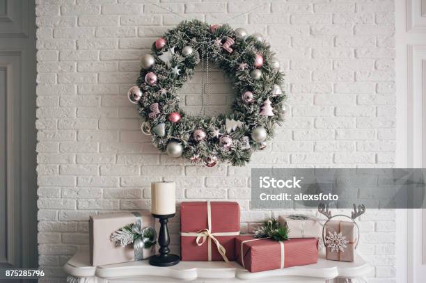 A Traditional Bright Christmas Wreath Hanging Over The Fireplace On A White Brick Wall And Packaged Gifts Are Stacked On A Fireplace With Candles Stock Photo - Download Image Now