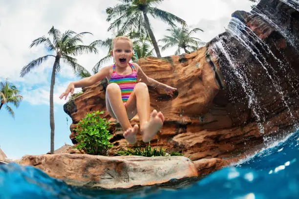Funny child learn to swim with fun. Girl jumping high with splashes into water pool under waterfall. Healthy lifestyle, kids water sport activity, swimming lesson with parents on family beach vacation