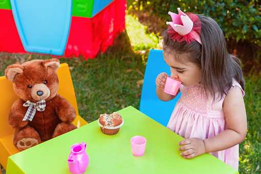 Baby toddler girl playing in outdoor tea party drinking from cup with best friend Teddy Bear sitting at table. Pink dress and queen or princess crown