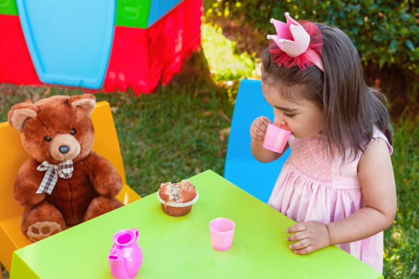 Baby toddler girl playing in outdoor tea party drinking from cup with best friend Teddy Bear sitting at table. Baby toddler girl playing in outdoor tea party drinking from cup with best friend Teddy Bear sitting at table. Pink dress and queen or princess crown kids play house stock pictures, royalty-free photos & images