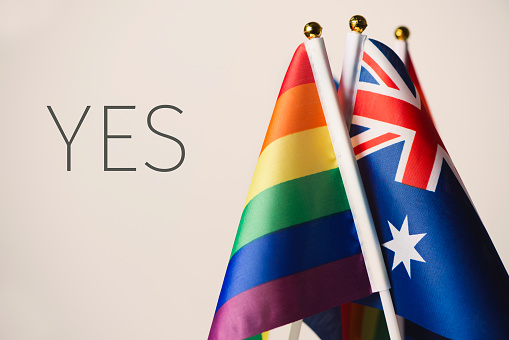 closeup of an australian flag and some rainbow flags, and the word yes, depicting the support to the same-sex union in Australia, against an off-white background