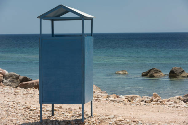 A simple blue painted changing booth or changing room at a rocky Greek Island beach in summer. In the background are the Aegean Sea, the horizon and the clear blue summer skies.