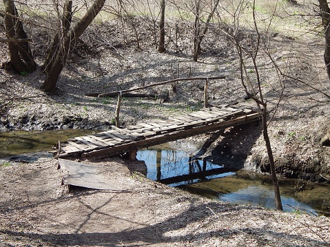 Old, rickety, wooden, pedestrian bridge. The bridge across the stream in spring forest. Photo.