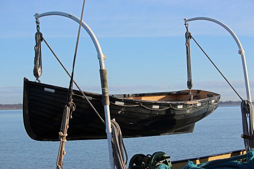 Black painted wooden lifeboat on davits. Background of river with bank in the distance and hazy blue sky.
