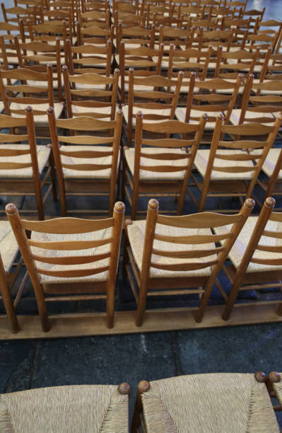 Multiple vintage wooden chairs with padded seats are neatly arranged in rows inside a hall. The worn wood and orderly alignment create a nostalgic and structured atmosphere. The image conveys themes of tradition, gathering, and preparation for an event or meeting.