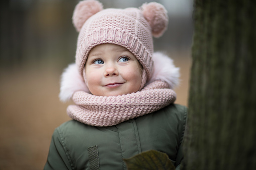 Pretty toddler girl ina rose scarf and hat is smiling looking up right leaning against the tree in autumn in Tavricheskiy Sad in StPete, Russia