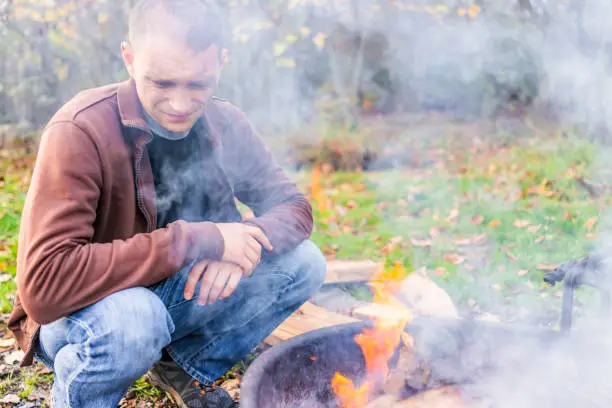 Young dirty gritty man with face covered in grey ash blowing campfire bonfire flame in fire pit frill by logs and autumn forest, lots of smoke