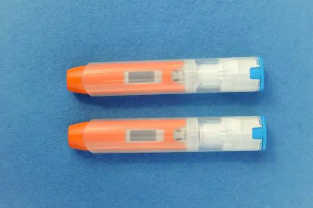 Two unused anaphylaxis Auto injectors on brightly coloured background. Still has safety cap in place. Clear window contains epinephrine.