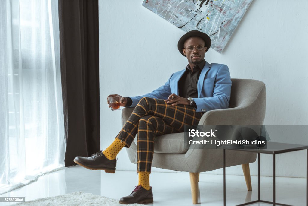 man with cigar and alcohol stylish african american man in hat, checkered pants and blue jacket with cigar and glass of cognac sitting in armchair in modern office Men Stock Photo