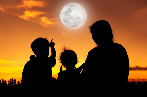 Silhouette back view of family sitting and relaxing together. Boy point to full moon on gold sky background. Happy family spending time together. The moon taken with my own camera, no NASA images used