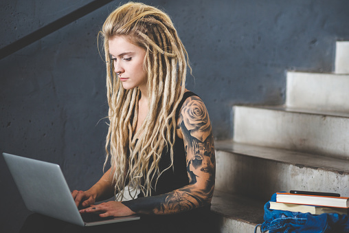 Portrait of young tattooed woman with blond dreadlocks sitting on floor and using notebook