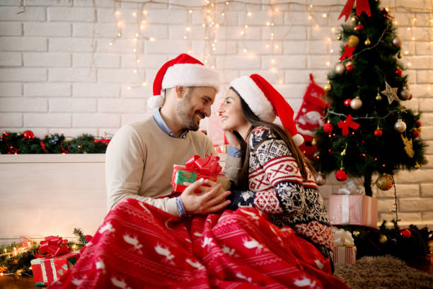 istock Cheerful happy young love couple sitting on a carpet with a gift while cuddling and hugging with Santa hats for Christmas holidays. 875722738