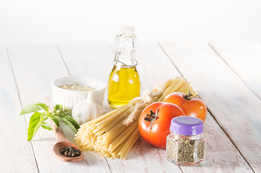Raw spaghetti, on a white wooden table, with tomatoes, garlic, black pepper, olive oil and another ingredients used to cook a pasta.