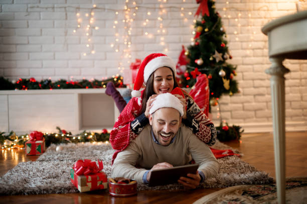 liebenswert verspielten paar genießt weihnachtsferien während girl auf freund rücken liegend und mit nikolausmütze und ein mann liegt auf einem teppich während der suche in einer tablette mit keksen in der nähe spielen. - couple loving lying on back carpet stock-fotos und bilder