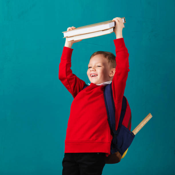 alegre pensativo escuela niño en la escuela con la mochila y la gran pila de libros contra la pared azul. mirando a cámara. concepto de escuela. regreso a la escuela - little boys measuring expressing positivity intelligence fotografías e imágenes de stock