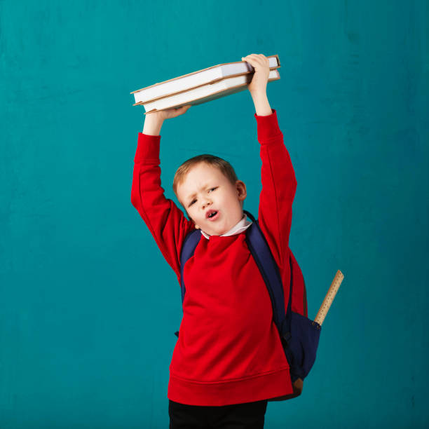 allegro ragazzino premuroso in uniforme scolastica con zaino e grande mucchio di libri in piedi contro il muro blu. guardando la telecamera. concetto scolastico. ritorno a scuola - little boys measuring expressing positivity intelligence foto e immagini stock