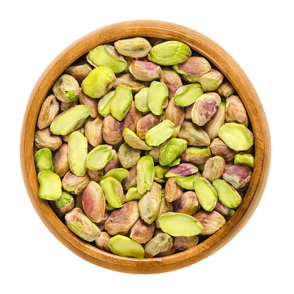 Shelled pistachio kernels in wooden bowl. Dried seeds and ripe fruits of Pistacia vera. Snack. Isolated macro food photo close up from above on white background.