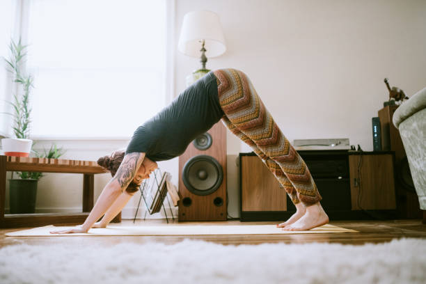 Young Adult Woman At Home Practicing Yoga A cheerful woman enjoys performing yoga stretches and exercise routines in her living room, the sun shining brightly through the windows. downward facing dog position stock pictures, royalty-free photos & images