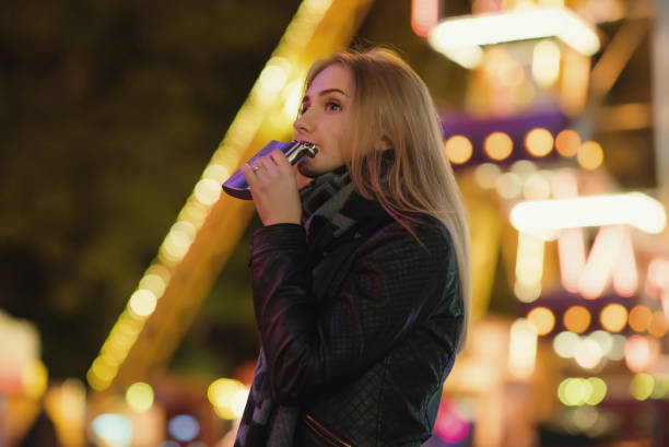 istock young beautiful woman alone drinks whiskey alcohol from flask. Outdoors at night in an amusement park. Night city background 875884724
