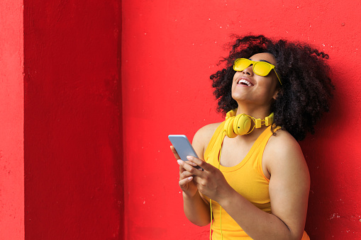 Smiling african woman with yellow sunglasses listening music on smart phone in front of bold colored background.