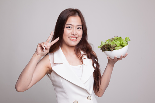 Healthy Asian woman show victory sign with salad on gray background