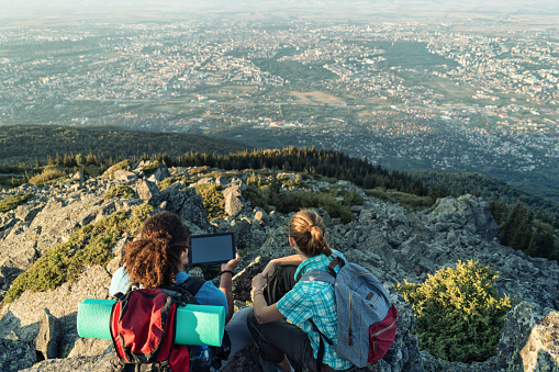 Rear view image of interracial couple relaxing on the rocky mountain top, holding digital tablet and looking at the valley.